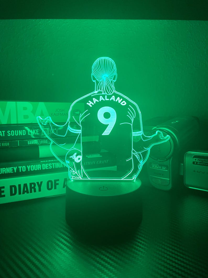 May include: A green-lit LED lamp featuring a soccer player in a meditative pose. The player's jersey displays "HAALAND" and the number "9". The lamp sits on a black base, with books and electronics in the background.