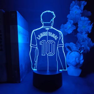 Puede incluir: Lámpara LED azul con una camiseta de fútbol con el nombre "LAMINE YAMAL" y el número "10" en la espalda. La lámpara está sobre una base negra, con un fondo oscuro e iluminación azul.