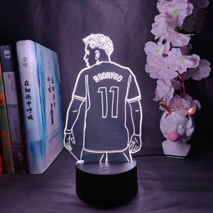 May include: A glowing LED lamp featuring a soccer player design. The player is wearing a jersey with the name "RODRYGO" and the number "11". The lamp sits on a black base and is surrounded by books and decorative flowers.