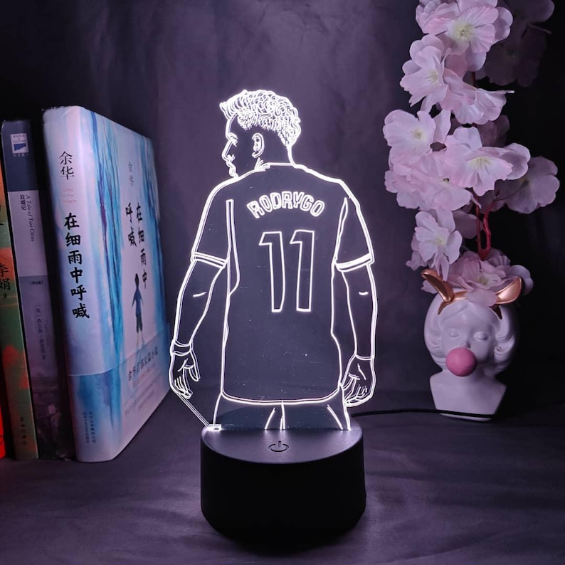 May include: A glowing LED lamp featuring a soccer player design. The player is wearing a jersey with the name "RODRYGO" and the number "11". The lamp sits on a black base and is surrounded by books and decorative flowers.