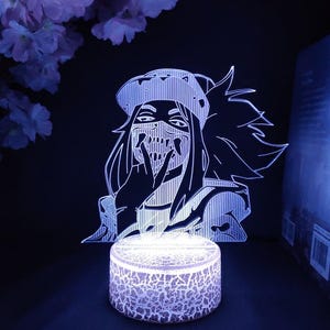 Puede incluir: Luz de noche LED brillante con un personaje de anime estilizado con m&aacute;scara y sombrero. El personaje est&aacute; representado en l&iacute;neas blancas sobre un fondo oscuro, con una base redonda y texturizada que emite una luz suave y fr&iacute;a. Dise&ntilde;o moderno y art&iacute;stico.