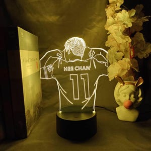 Peut inclure: Veilleuse LED en forme de maillot de football avec le nom "HEE CHAN" et le numéro "11". La lumière blanche repose sur un socle noir. Un livre et un vase décoratif sont visibles en arrière-plan.
