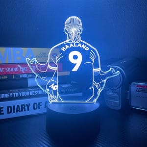 May include: A neon-style LED lamp featuring a soccer player's jersey with the name "HAALAND" and the number "9". The lamp is mounted on a black base and emits a blue glow. The design is a modern take on sports memorabilia.
