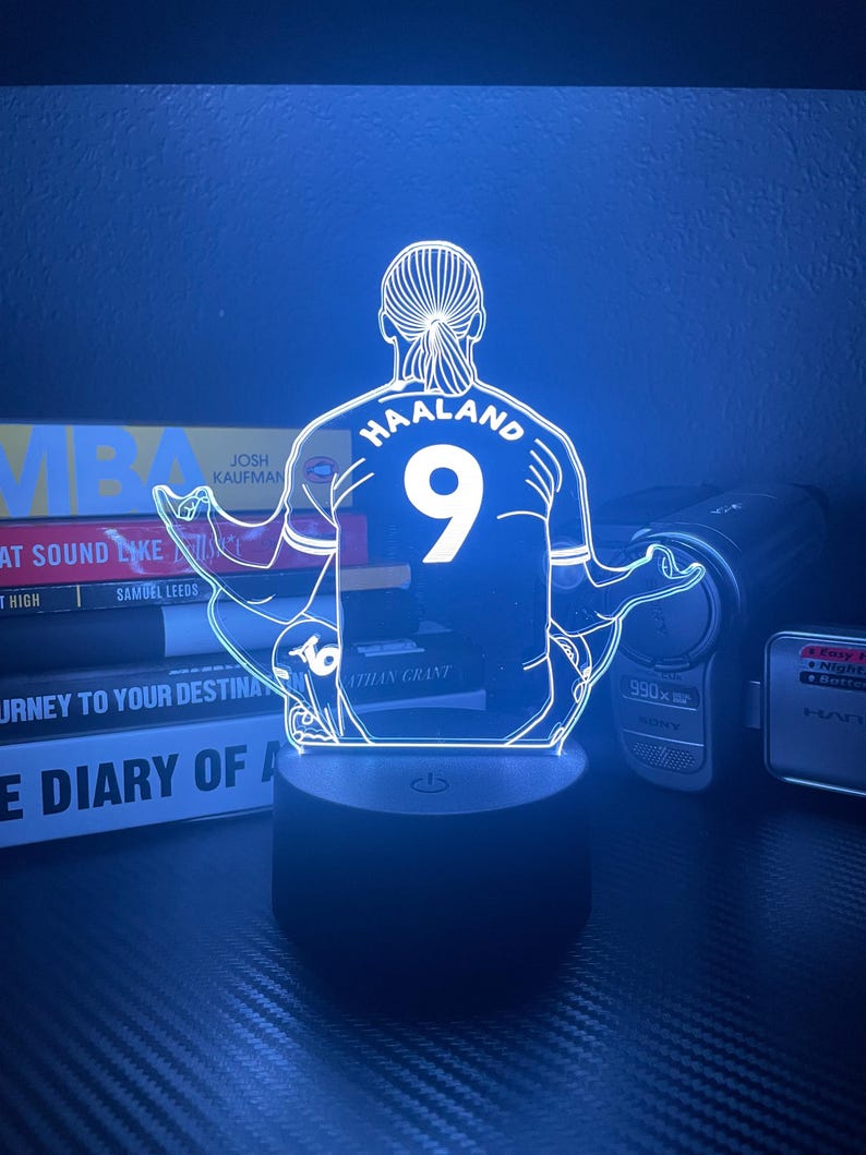 May include: A neon-style LED lamp featuring a soccer player's jersey with the name "HAALAND" and the number "9". The lamp is mounted on a black base and emits a blue glow. The design is a modern take on sports memorabilia.