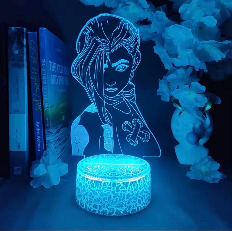 May include: A blue LED lamp featuring a character design, with a cracked-glass effect base. The lamp is next to books and decorative flowers. The books are titled "Call Me By Your Name" and "The Old Man and the Sea".
