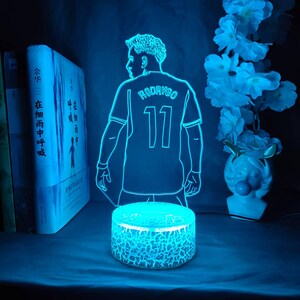 May include: A blue LED night light featuring a soccer player's silhouette, with the name "RODRYGO" and the number "11" on the jersey. The light sits on a cracked-glass-style base, with books and flowers in the background.