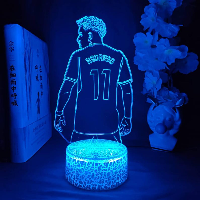May include: A blue LED night light featuring a soccer player in a jersey with the name "RODRYGO" and the number "11". The light is on a round base with a cracked glass effect, and is next to a book and a vase of flowers.