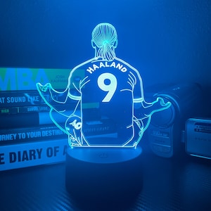 以下が含まれることがあります： サッカー選手の背中を模した青色のLEDランプ。背中には「HAALAND」の文字と「9」の番号が描かれています。ランプは黒い台座にあり、青く光っています。背景には本が見えます。