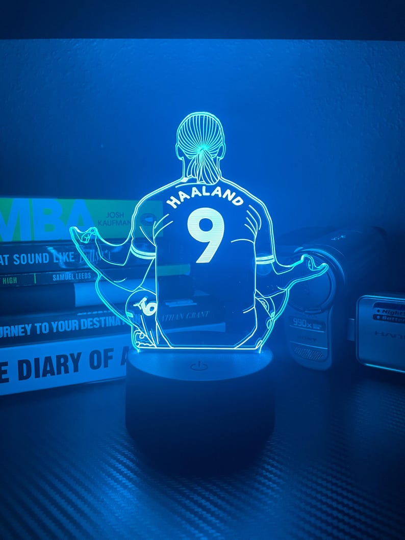 May include: A blue LED lamp featuring a stylized image of a soccer player's back, with the name "HAALAND" and the number "9" on the jersey. The lamp is on a black base and emits a blue glow. Books are visible in the background.