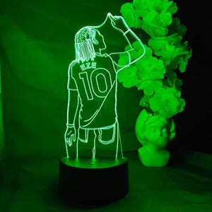 May include: A green LED lamp depicting a footballer in a jersey with "EZE" and the number "10". The lamp sits on a black base, with a green floral display in the background. The player is in a celebratory pose.