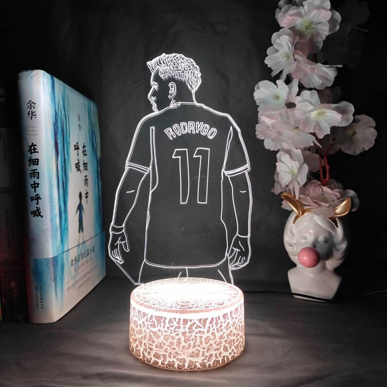 May include: A glowing LED lamp featuring a soccer player's jersey with the name "RODRYGO" and the number "11". The lamp is illuminated by a round, textured base. A book and decorative flowers are in the background.