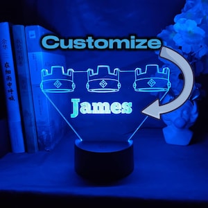 Puede incluir: Luz nocturna LED azul con el nombre "James" en blanco. El diseño presenta tres contornos de corona y la palabra "Personalizar". La luz se encuentra sobre una base negra, creando una decoración personalizada y brillante.