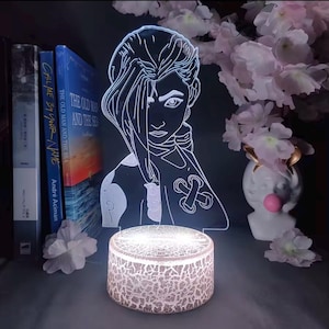 May include: A glowing LED lamp featuring a stylized illustration of a character with long hair. The lamp sits on a textured, round base. Books and decorative flowers are in the background. The lamp's design is a white outline on a clear surface.