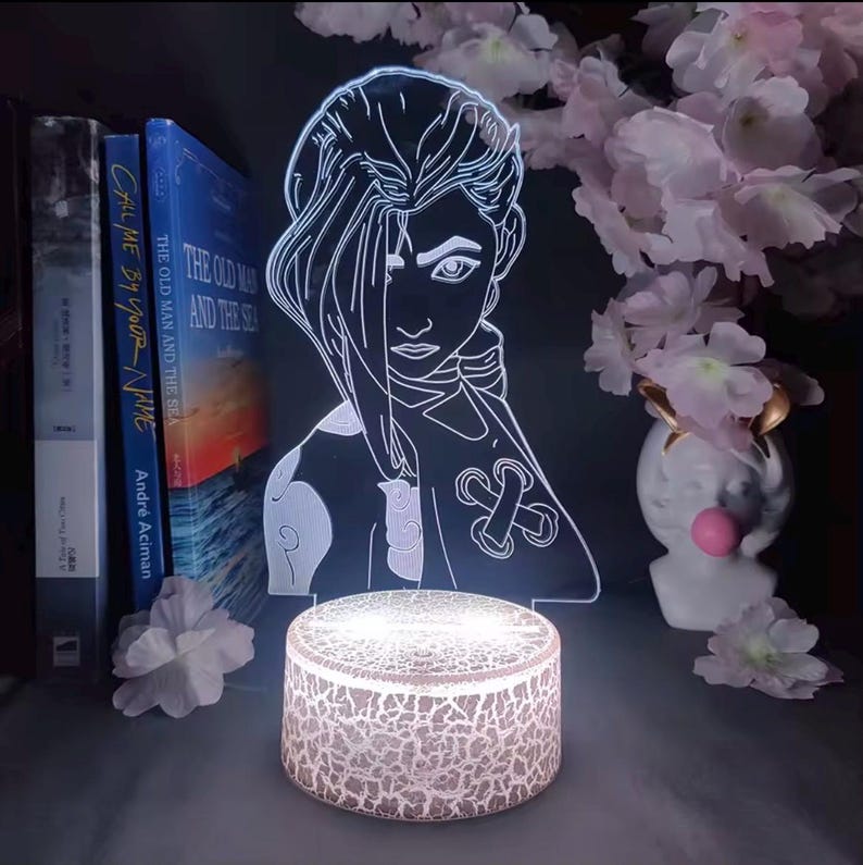 May include: A glowing LED lamp featuring a stylized illustration of a character with long hair. The lamp sits on a textured, round base. Books and decorative flowers are in the background. The lamp's design is a white outline on a clear surface.