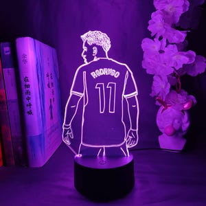May include: A purple LED lamp featuring a soccer player's jersey with the name "RODRYGO" and the number "11". The lamp is on a black base, with a silhouette design. The background includes books and flowers.