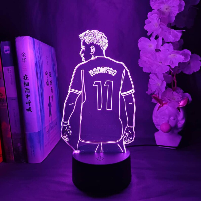 May include: A purple LED lamp featuring a soccer player's jersey with the name "RODRYGO" and the number "11". The lamp is on a black base, with a silhouette design. The background includes books and flowers.