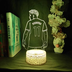 May include: A glowing LED night light featuring a soccer player's jersey with the name "RODRYGO" and the number "11". The light is a white outline on a green background, with a textured, cylindrical base. Books and a decorative vase are in the background.
