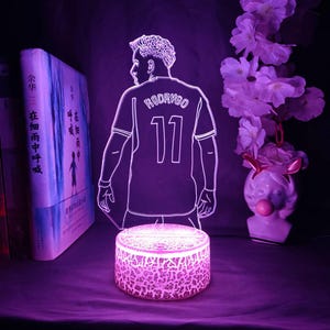 May include: A glowing purple LED lamp featuring a soccer player's silhouette, with the name "RODRYGO" and the number "11" on the jersey. The lamp sits on a cracked-glass-style base, with a book and decorative flowers in the background.