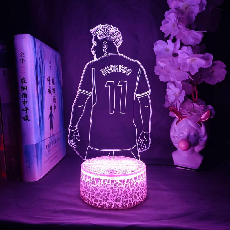 May include: A glowing purple LED lamp featuring a soccer player's silhouette, with the name "RODRYGO" and the number "11" on the jersey. The lamp sits on a cracked-glass-style base, with a book and decorative flowers in the background.