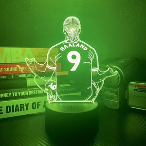 May include: A green-lit LED lamp featuring a soccer player's silhouette, with the name "HAALAND" and the number "9" on the jersey. The lamp sits on a black base, illuminating the figure. Books and electronics are in the background.
