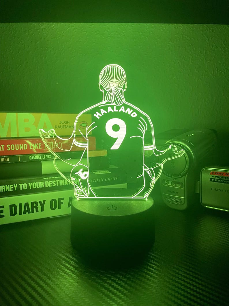 May include: A green-lit LED lamp featuring a soccer player's silhouette, with the name "HAALAND" and the number "9" on the jersey. The lamp sits on a black base, illuminating the figure. Books and electronics are in the background.