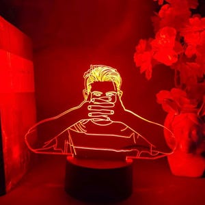 Può includere: Lampada a LED rossa raffigurante una persona che si copre la bocca con le mani. La lampada ha una base nera e un contorno luminoso del viso, dei capelli e della camicia della persona. Lo sfondo è rosso scuro, con fiori rossi.