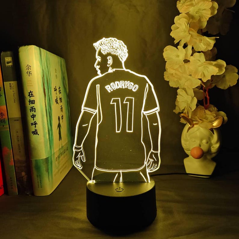 May include: A glowing LED lamp featuring a soccer player's silhouette, wearing a jersey with "RODRYGO" and the number "11". The lamp has a black base and is illuminated with a warm, inviting light. Decorative items are in the background.
