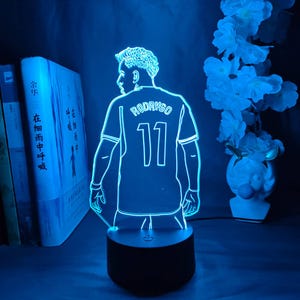 May include: A blue LED night light featuring a soccer player's jersey with the name "RODRYGO" and the number "11". The light is on a black base, with a silhouette of the player. The background includes books and white flowers.