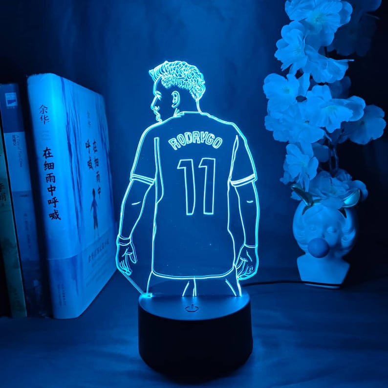 May include: A blue LED night light featuring a soccer player's jersey with the name "RODRYGO" and the number "11". The light is on a black base, with a silhouette of the player. The background includes books and white flowers.