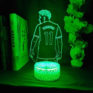 May include: A green LED night light featuring a soccer player in a jersey with the name "RODRYGO" and the number "11". The light has a cracked-glass effect base and is surrounded by books and decorative flowers.