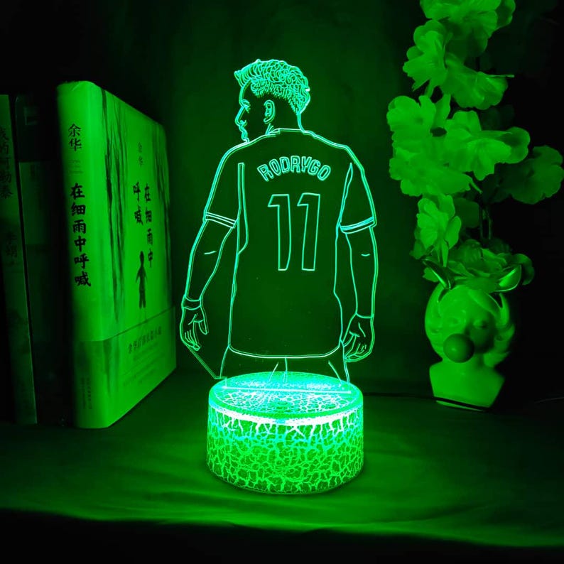 May include: A green LED night light featuring a soccer player in a jersey with the name "RODRYGO" and the number "11". The light has a cracked-glass effect base and is surrounded by books and decorative flowers.