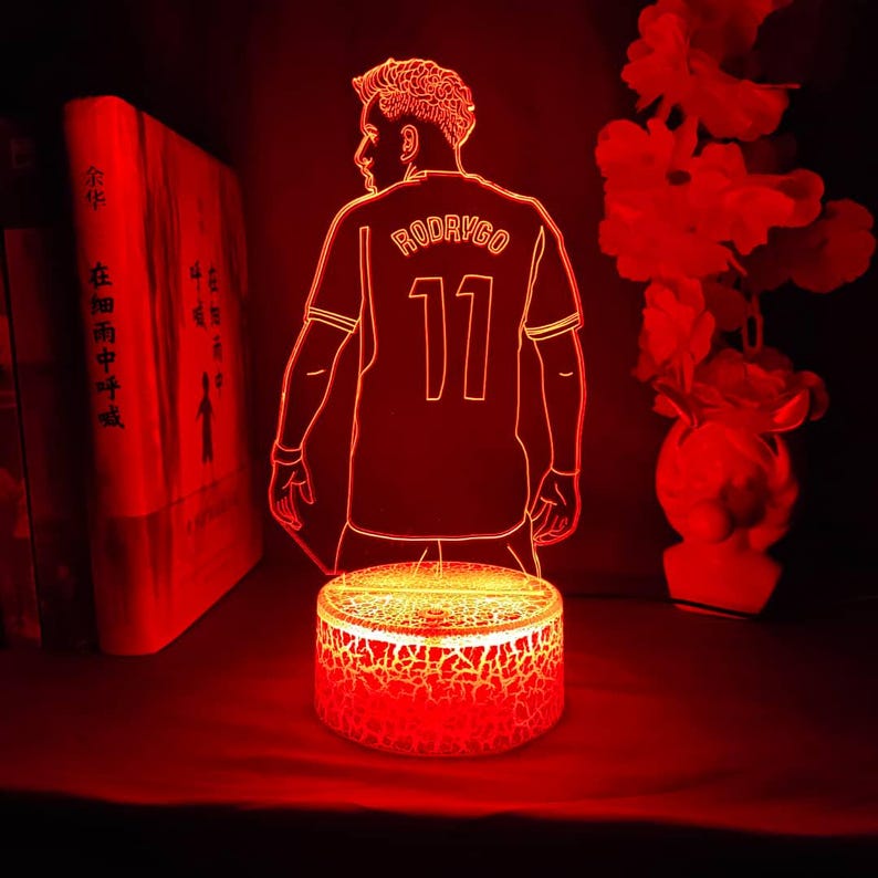 May include: A red LED night light featuring a soccer player in a jersey with the name "RODRYGO" and the number "11". The light is on a cracked-effect base, with a red glow illuminating the figure and the surrounding area.