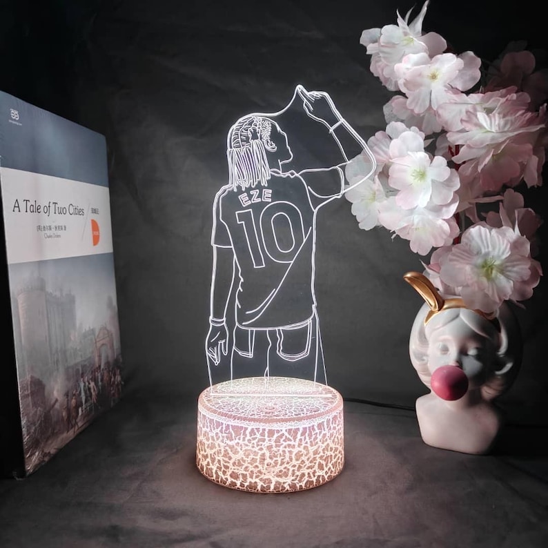 May include: An illuminated LED lamp with a soccer player design, displaying the name "EZE" and the number "10" on the jersey. The lamp has a round base with a crackle pattern. A book titled "A Tale of Two Cities" is visible.