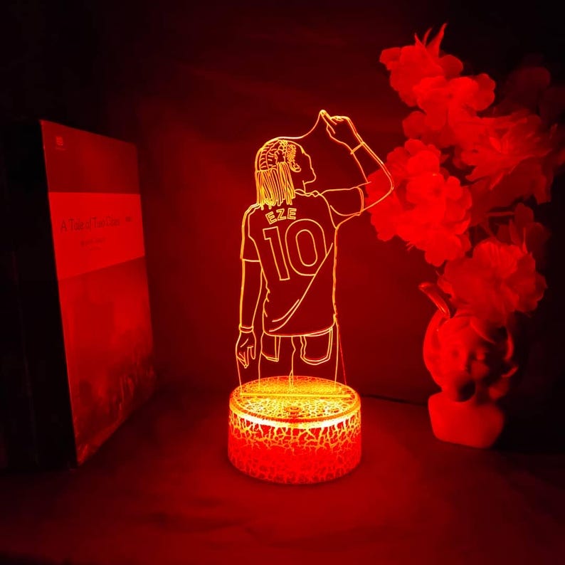 May include: A red LED lamp depicting a soccer player wearing a jersey with "EZE" and the number "10". The lamp has a round base with a cracked design. The background is dark, with a book and decorative flowers.