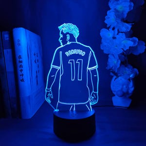 May include: A blue LED lamp featuring a soccer player's jersey with the name "RODRYGO" and the number "11" illuminated. The lamp has a black base and is surrounded by books and flowers.