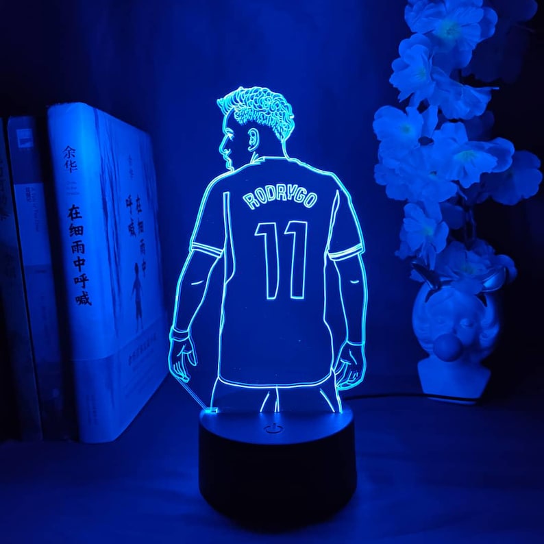 May include: A blue LED lamp featuring a soccer player's jersey with the name "RODRYGO" and the number "11" illuminated. The lamp has a black base and is surrounded by books and flowers.