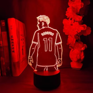 May include: A red LED lamp featuring a soccer player in a jersey with the name "RODRYGO" and the number "11". The lamp is on a black base, with a red glow illuminating the figure and surrounding area. Books and flowers are in the background.
