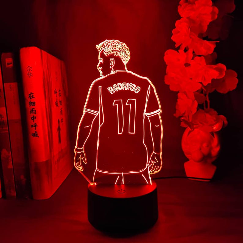 May include: A red LED lamp featuring a soccer player in a jersey with the name "RODRYGO" and the number "11". The lamp is on a black base, with a red glow illuminating the figure and surrounding area. Books and flowers are in the background.