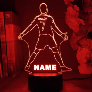 May include: A red LED night light featuring a laser-cut acrylic silhouette of a soccer player in a goal celebration pose. The design includes the text RONALDO, the number 7, and a placeholder for a name, all mounted on a black circular base.