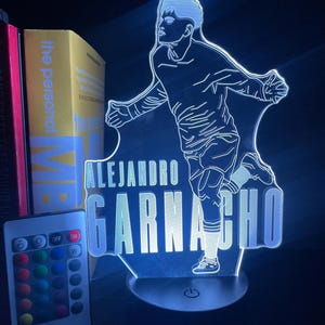 Puede incluir: Lámpara LED azul con la silueta de un jugador de fútbol y el nombre "ALEJANDRO GARNACHO". La lámpara incluye un mando a distancia para ajustar el color. La lámpara está sobre una superficie negra.