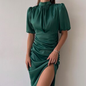 May include: Emerald green satin dress with a high neckline, short puff sleeves, and a thigh-high slit. The dress features ruching on the side and a flowing skirt. A sophisticated formal wear option.