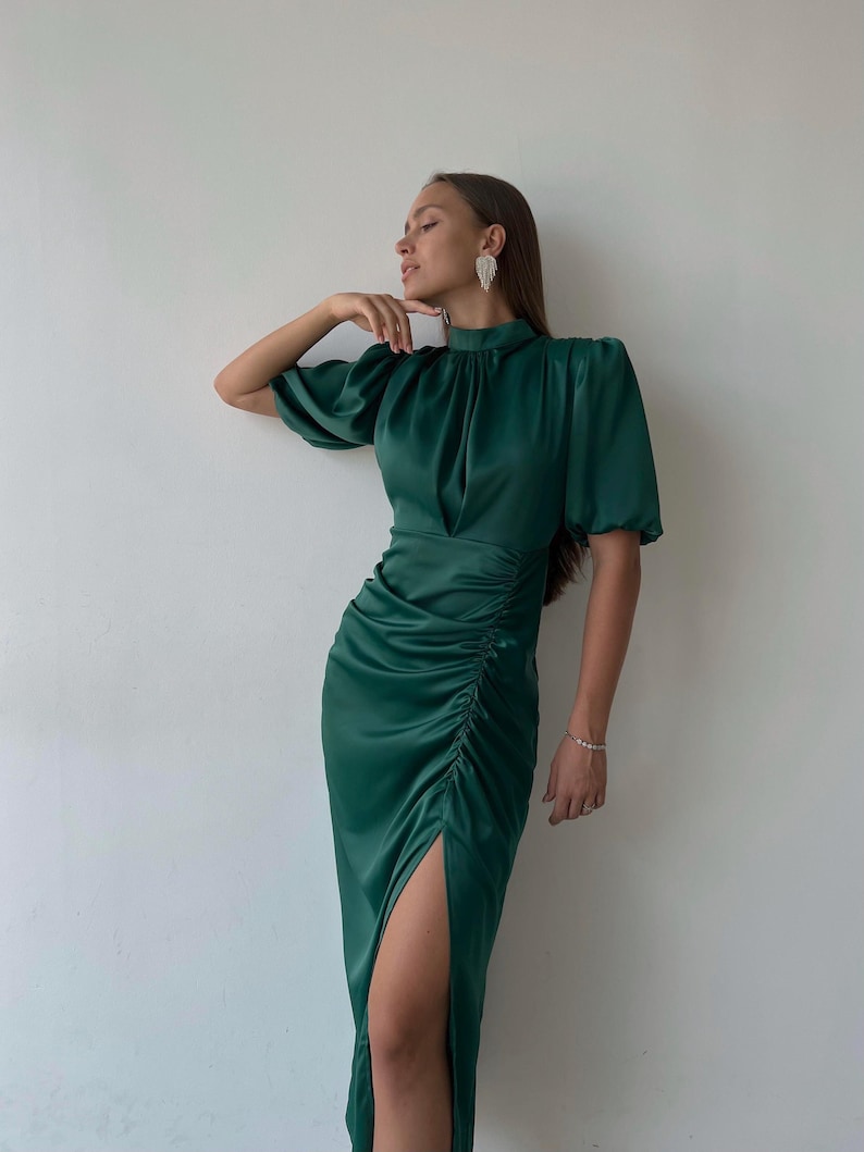 May include: A dark green satin dress with a high neckline and short sleeves. The dress features a ruched detail on the side and a thigh-high slit. The dress is a solid color and appears to be made of a smooth, flowing fabric.