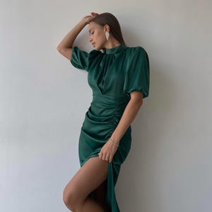 May include: A dark green satin dress with short, puffed sleeves and a high neckline. The dress features ruching on the side and a thigh-high slit. The model is wearing silver earrings and heels.