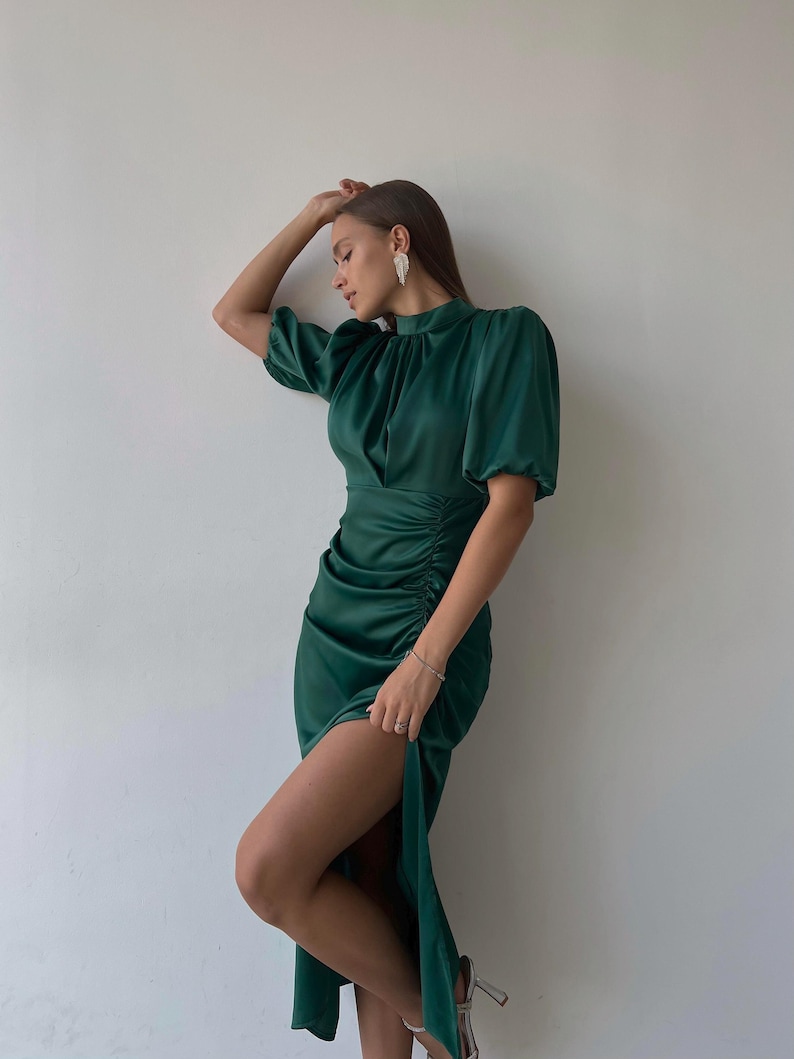 May include: A dark green satin dress with short, puffed sleeves and a high neckline. The dress features ruching on the side and a thigh-high slit. The model is wearing silver earrings and heels.