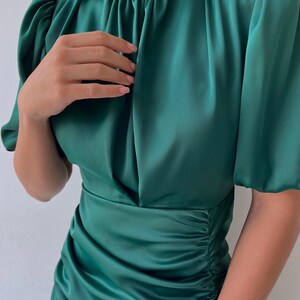 May include: A dark green satin dress with a high neckline and short, puffed sleeves. The dress features ruching at the waist and a flowing skirt, creating a flattering silhouette. The fabric has a subtle sheen, enhancing the overall elegance of the garment.
