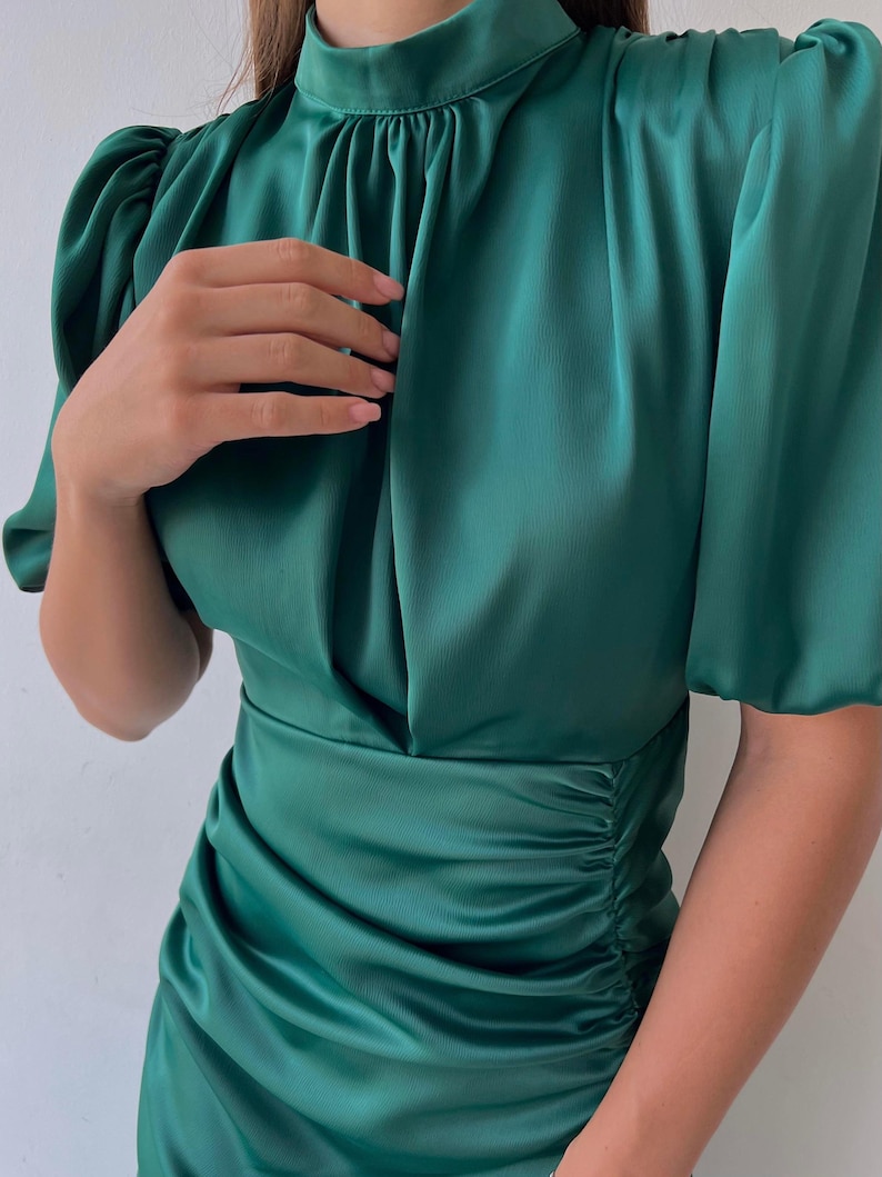 May include: A dark green satin dress with a high neckline and short, puffed sleeves. The dress features ruching at the waist and a flowing skirt, creating a flattering silhouette. The fabric has a subtle sheen, enhancing the overall elegance of the garment.