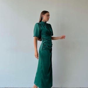 May include: A long, emerald green dress with short, puffed sleeves and a high neckline. The dress features a ruched detail on the side and is paired with silver high-heeled sandals. The dress is made of a shiny, satin-like material.