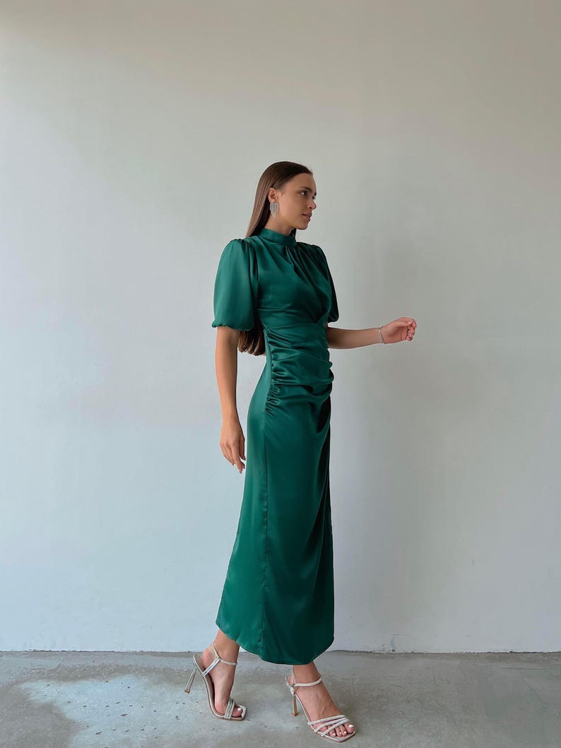 May include: A long, emerald green dress with short, puffed sleeves and a high neckline. The dress features a ruched detail on the side and is paired with silver high-heeled sandals. The dress is made of a shiny, satin-like material.
