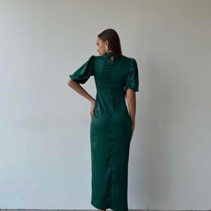 May include: A dark green satin dress with short, puffed sleeves and a high neckline. The dress has a fitted bodice and a long, straight skirt. The model is wearing silver heels.