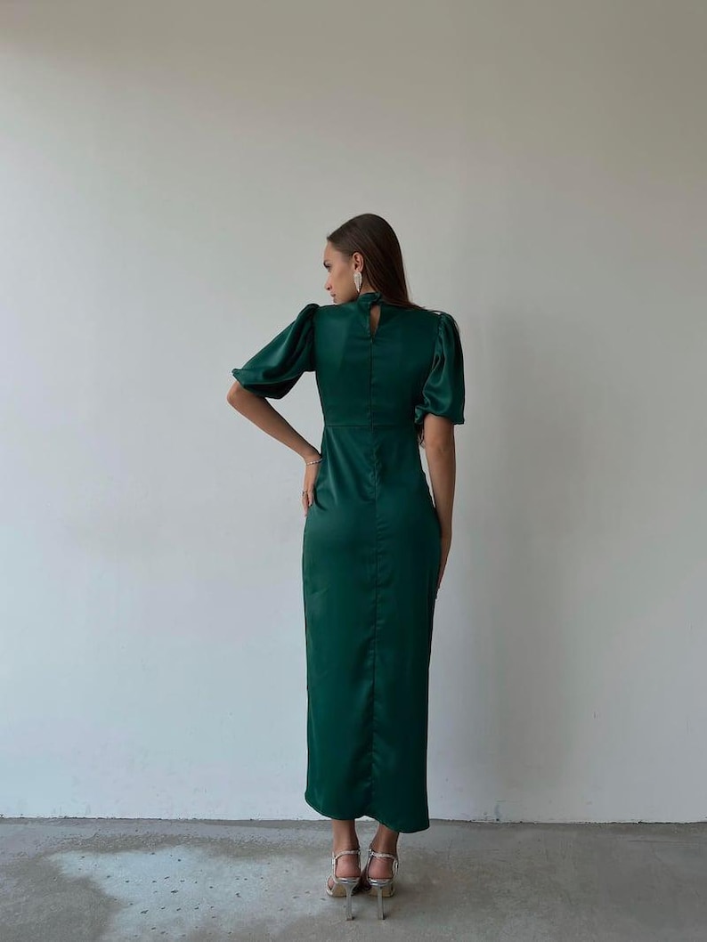 May include: A dark green satin dress with short, puffed sleeves and a high neckline. The dress has a fitted bodice and a long, straight skirt. The model is wearing silver heels.
